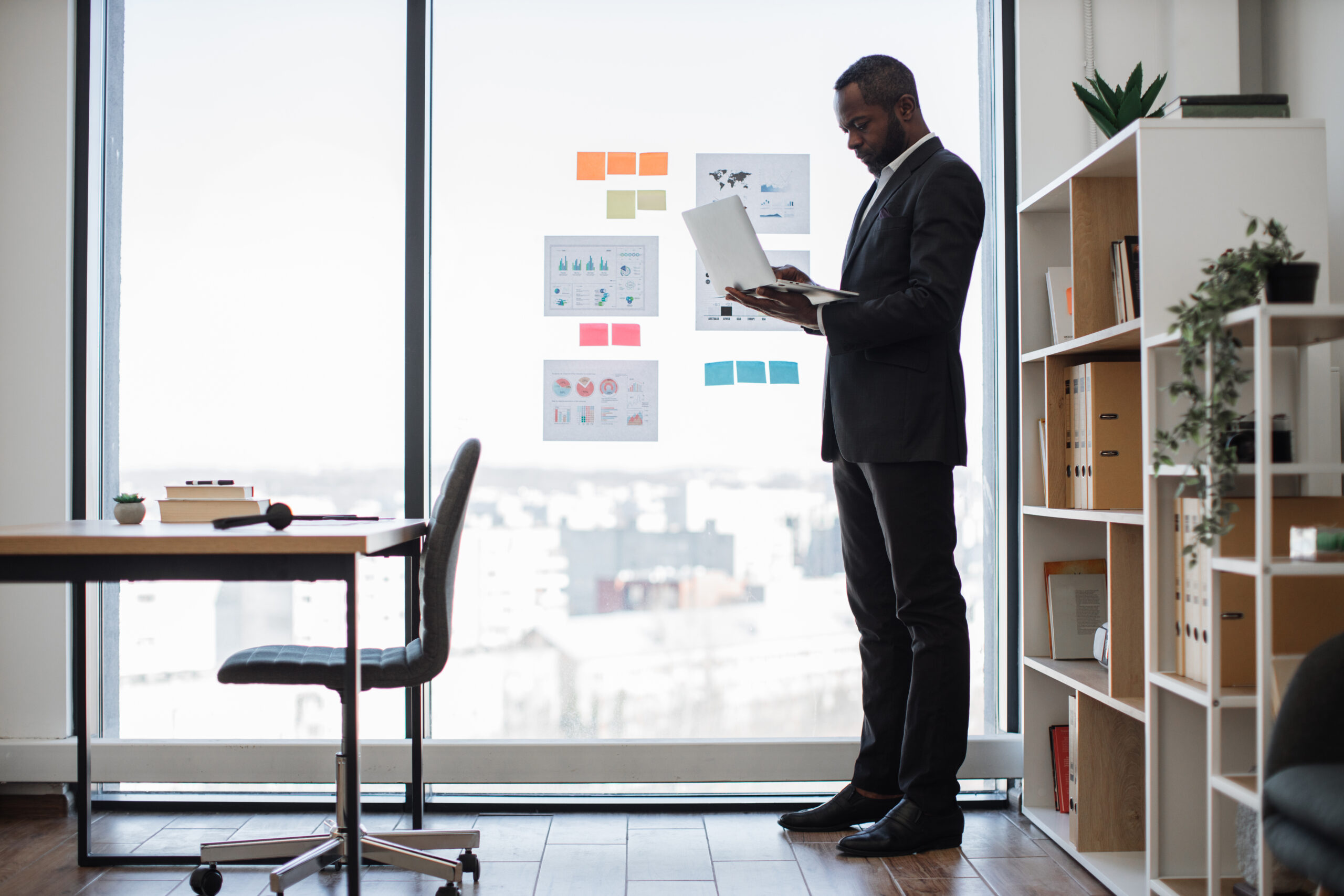 Stylish multicultural male in business suit standing in creative workplace with digital device in hands. Executive manager planning investment strategy using laptop and data visualization materials.